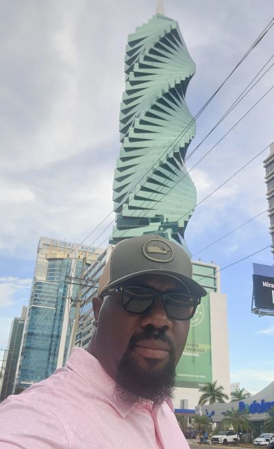 Man wearing glasses and cap stands in front of a twisting modern skyscraper.
