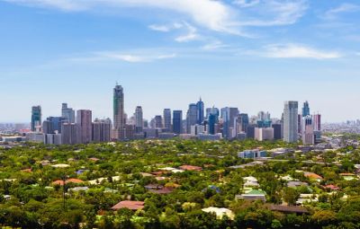 City skyline with lush green trees in the foreground under a blue sky.