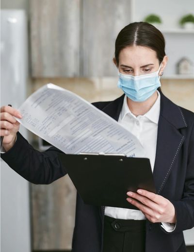 A woman wearing a mask reviews documents on a clipboard indoors.