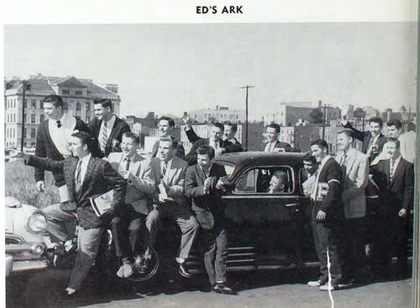 A group of young men dressed in suits gather around a classic car in a city setting.
