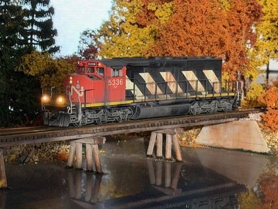 Model train crossing a small bridge over reflective water with autumn trees.