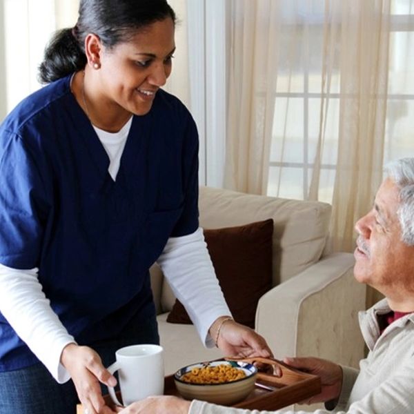A caregiver serving food and drink to an elderly man at home.