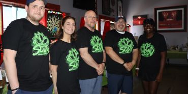 Group of five people wearing matching black shirts with a green logo indoors.