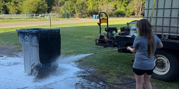 Person pressure washing a large trash bin outdoors.