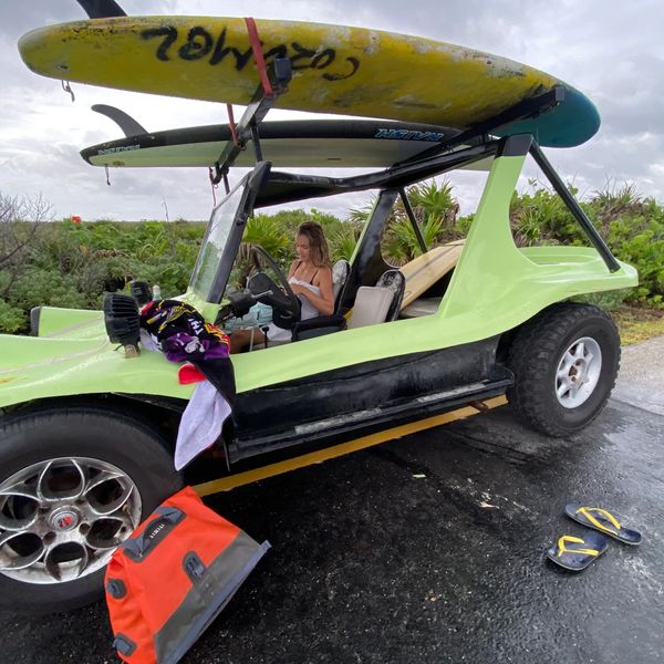A woman sits in a green dune buggy with surfboards on top.