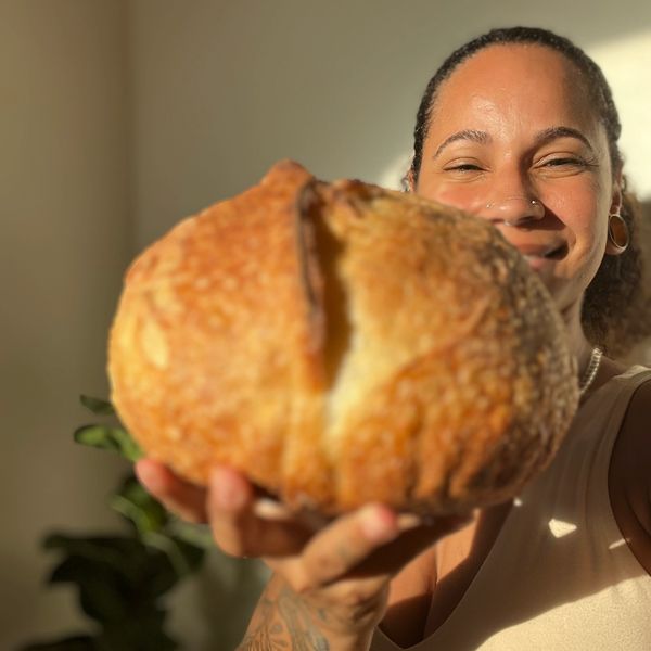 A smiling woman holding a loaf of bread towards the camera.