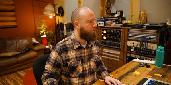 Callum Edwards  in plaid flannel shirt at wooden desk with computer in studio filled with audio equi