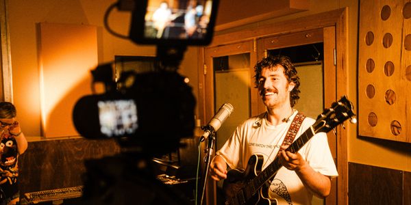 The Grogans singer playing black electric guitar and singing into mic, filmed in warm-lit studio