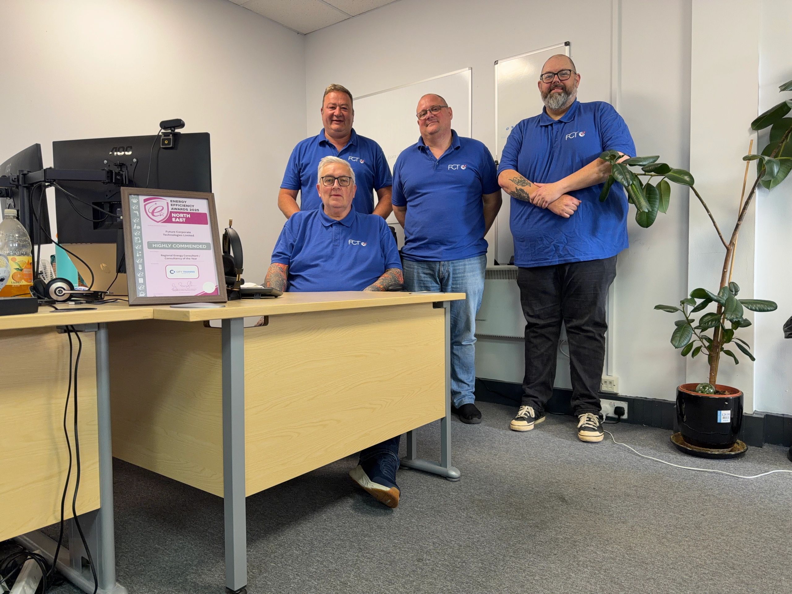 Four men in blue shirts posing in an office with an award on the desk.