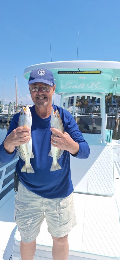 Man on boat holding two fish, smiling under clear blue sky.