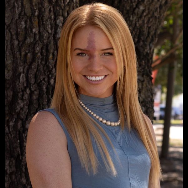 Smiling woman with long blonde hair and pearl necklace outdoors.