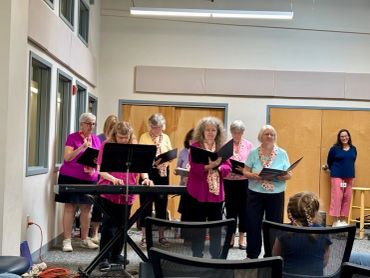 A group of elderly women singing in a room, with a keyboard in front.