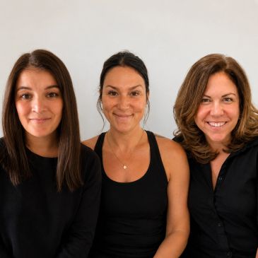 Three women smiling together against a plain background.
