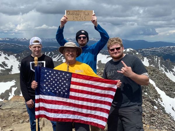 Four hikers celebrate reaching Quandary Peak summit, holding an American flag and a sign with the elevation.