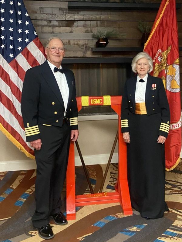 Steve and his wife in formal military attire pose beside a USMC stand with American and Marine flags