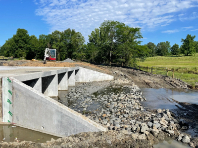 Newly constructed concrete bridge over a small stream with rocks and green trees around.