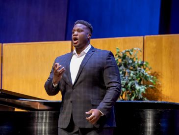 A man passionately singing on stage beside a piano.