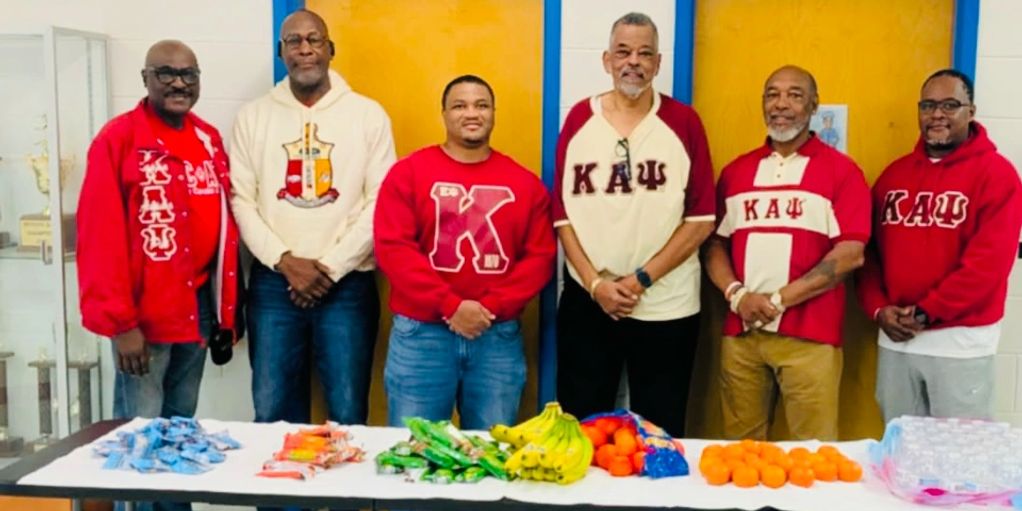 Six men standing behind a table with snacks, wearing fraternity apparel.