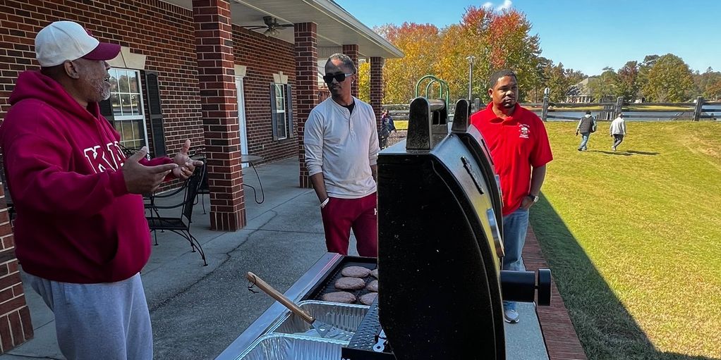 Three men chatting near a grill with burgers on it outdoors on a sunny day.