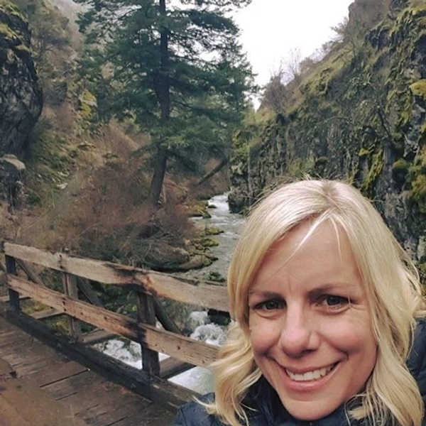 Smiling woman on a wooden bridge with a forested river gorge in the background.