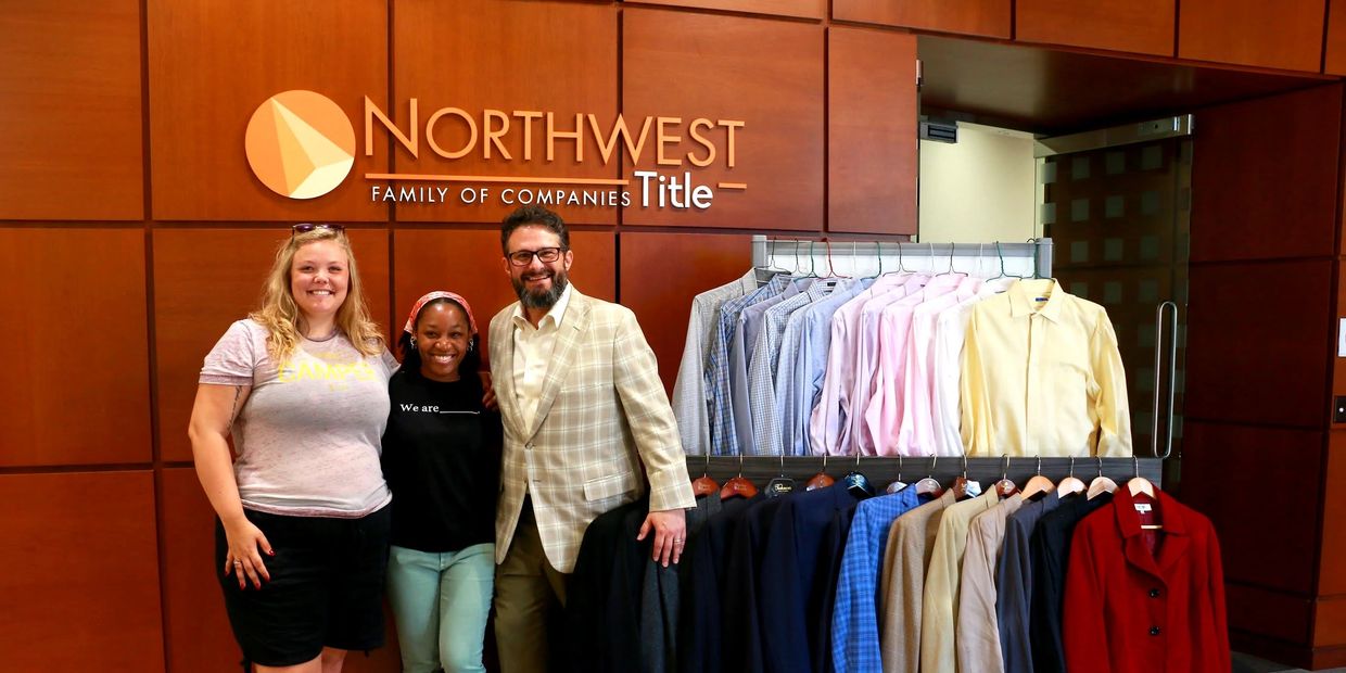 Three people stand smiling in front of a rack of shirts at Northwest Title office.