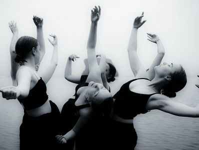 A group of dancers in black outfits performing expressive movements by the water.