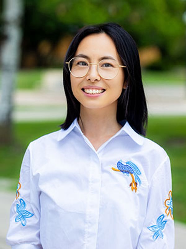 Smiling woman wearing glasses and a white shirt with blue and orange embroidery outdoors.