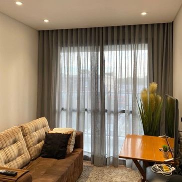 Sala com cortina wave, sofá marrom com almofadas, uma planta decorativa, mesa e televisão.