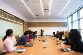 Diverse group of people seated around a conference table watching someone writing on a whiteboard. 