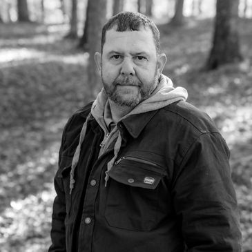 Bearded man in a jacket stands outdoors in a forest.