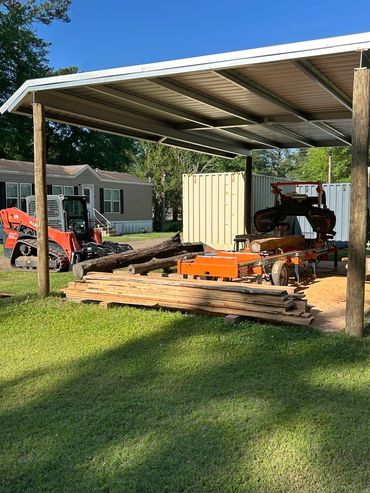 Woodworking setup with sawmill and tractor under a metal shed.