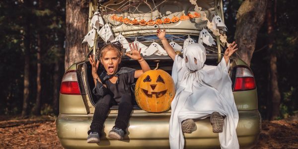 Two kids in Halloween costumes sitting on a car trunk with a pumpkin and ghost decorations.