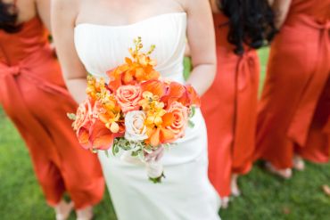 Bride holding vibrant orange and peach floral bouquet with bridesmaids in orange dresses.