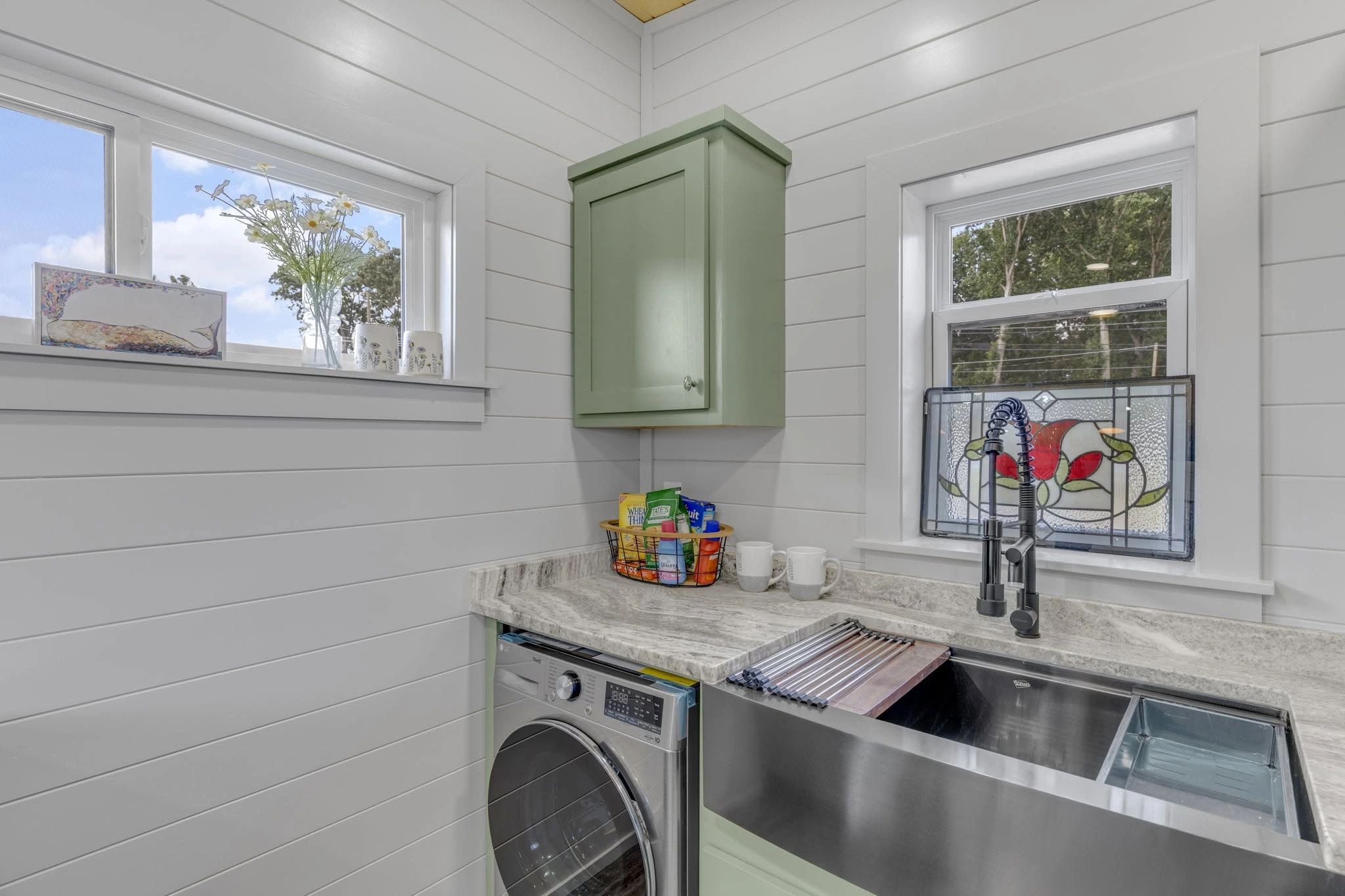 Modern laundry room with green cabinets and a stainless steel sink under windows.