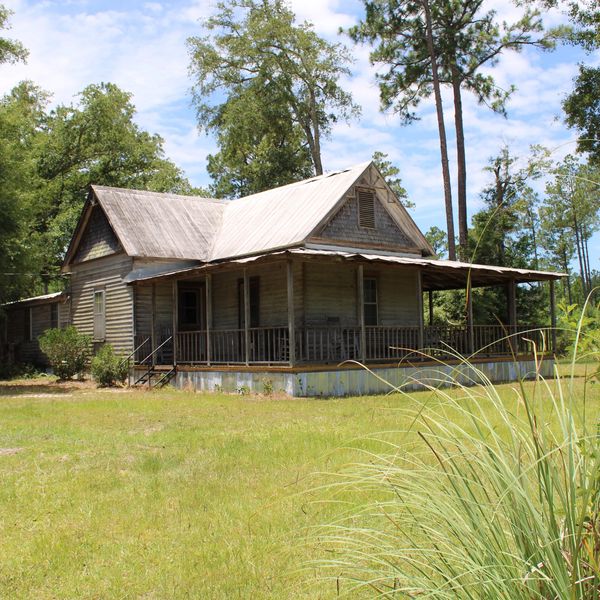 Old wooden house with a large porch surrounded by greenery.