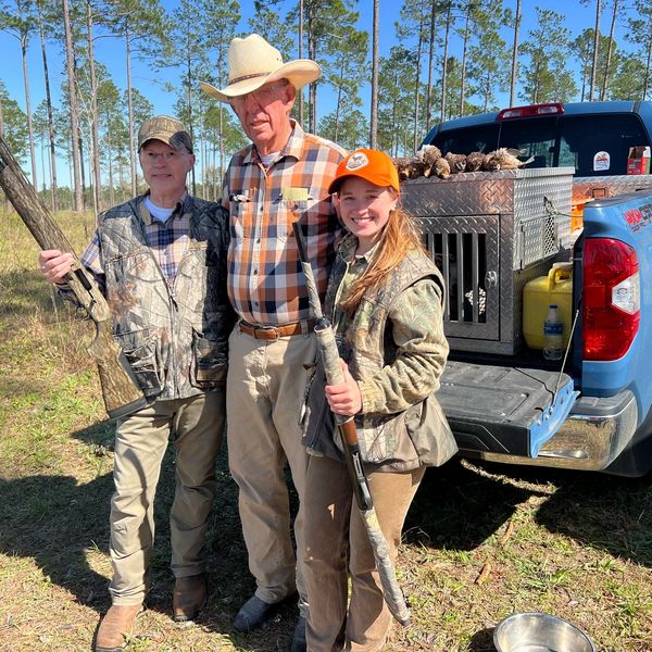 Three hunters standing in front of a blue pickup truck holding shotguns in a wooded area.