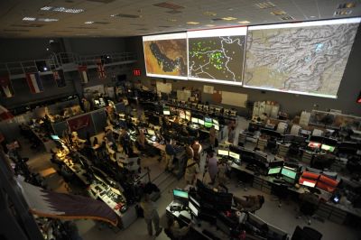 A busy control room with multiple monitors and large maps on the wall.