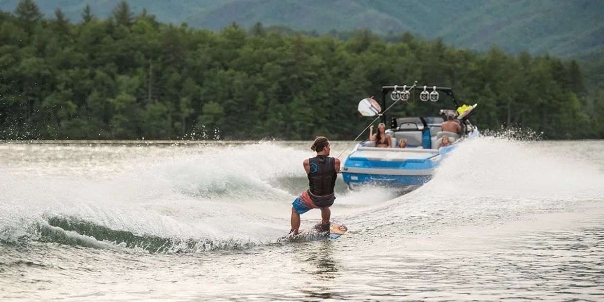 A man wakeboarding behind a boat on a lake surrounded by forested hills.