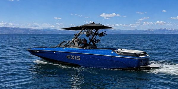 Blue Axis wakeboard boat cruising on a lake under a clear sky.