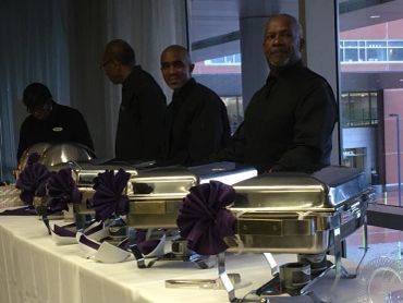 Four men dressed in black standing behind a buffet table with chafing dishes.