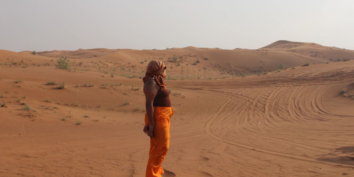 A person stands in the desert wearing bright orange pants and a headscarf, gazing at the dunes.