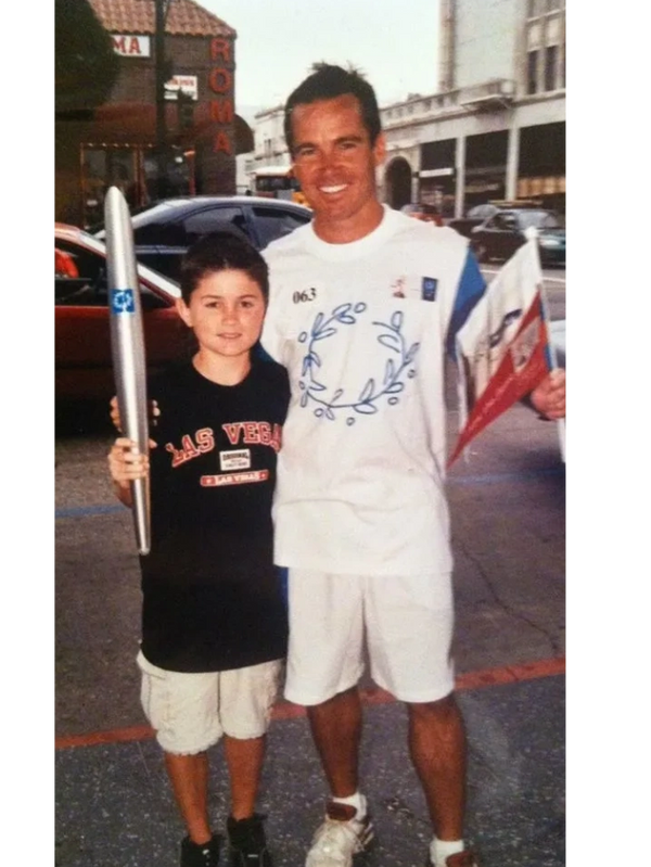 A boy and a man posing together with a torch and a flag, outdoors.