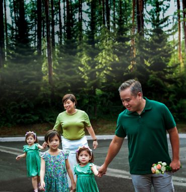 A family walks together in a forested area, holding hands and smiling.