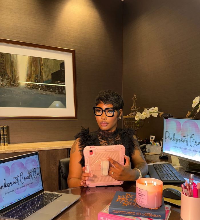 Woman in glasses holding a tablet at a desk with laptop and candle.