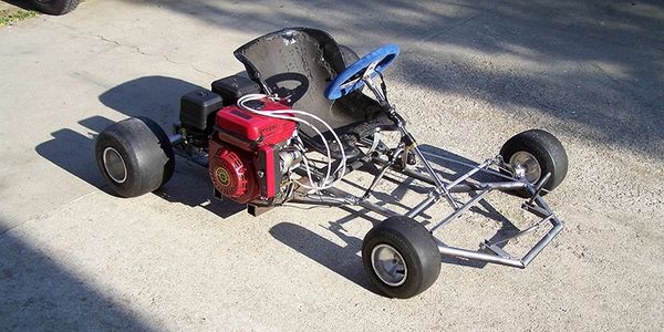 A homemade go-kart with a red engine and blue steering wheel on a concrete surface.