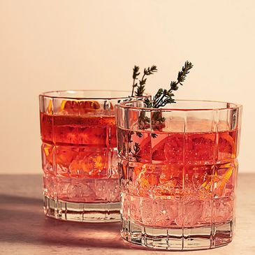 Two glasses of red cocktail garnished with herbs and ice.
