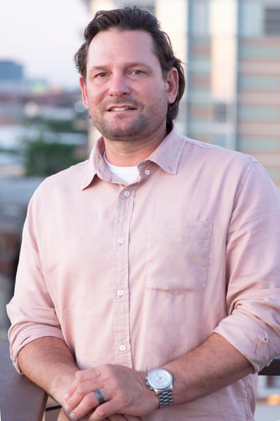 Man in a light pink shirt standing outdoors with a cityscape background.