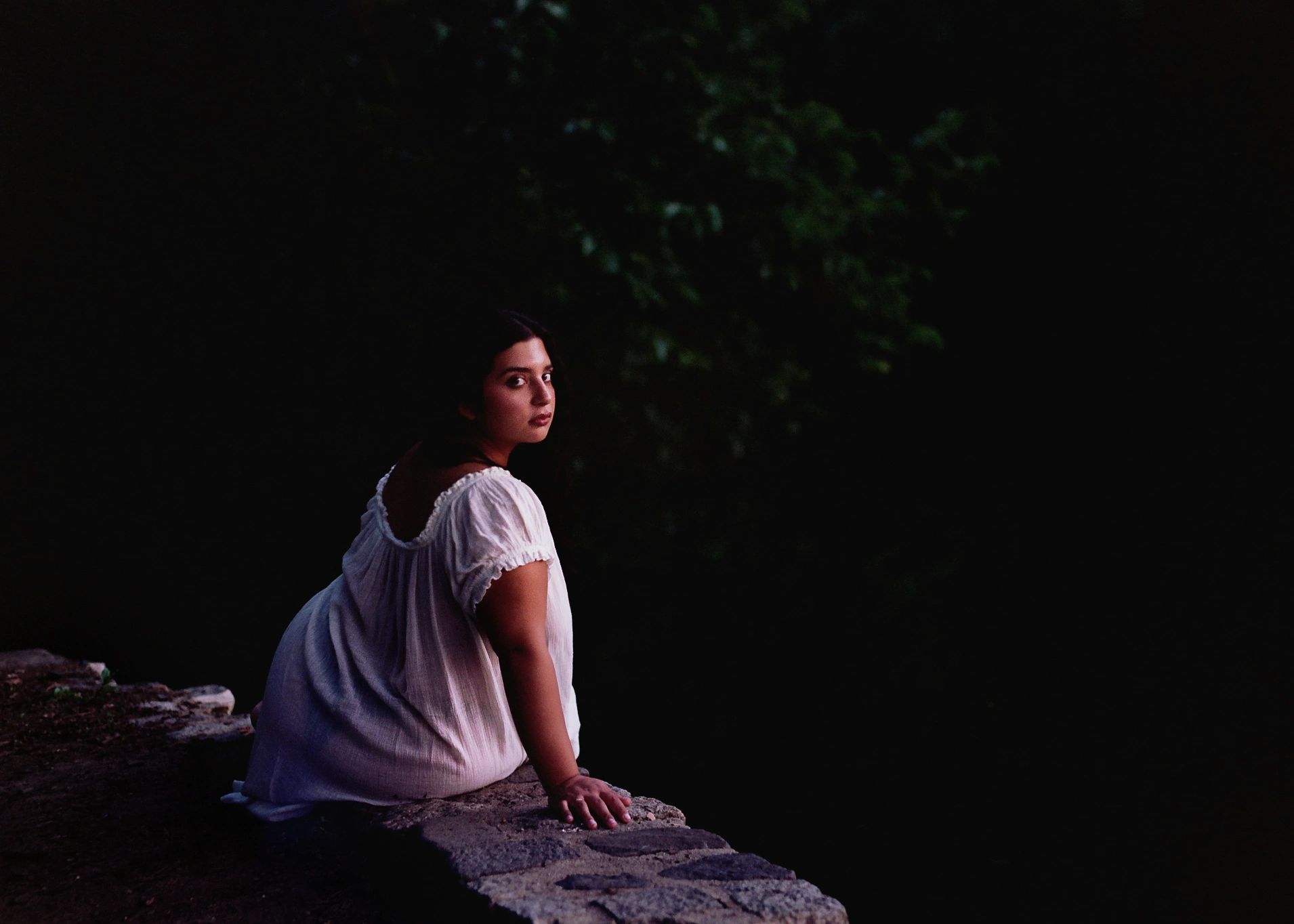 Woman in white dress sitting on stone ledge, looking back in dim light.