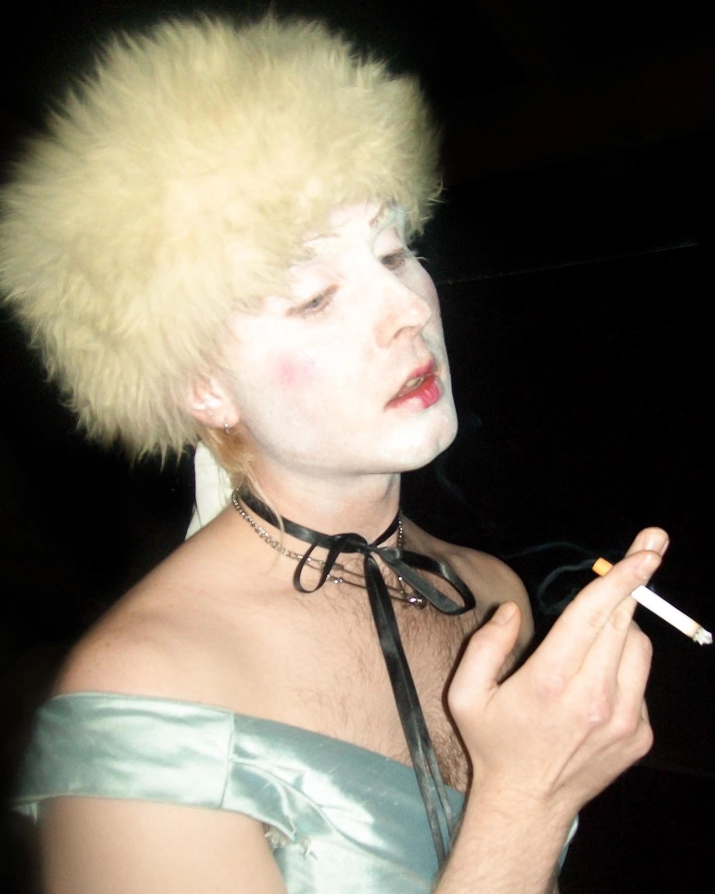 Person with white face makeup and fluffy hat smoking a cigarette.