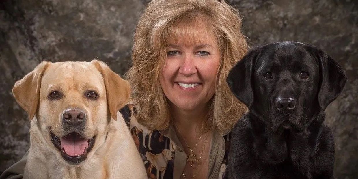 A smiling woman posing with two Labrador Retrievers, one yellow and one black.